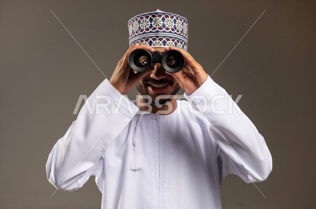 حب الاستكشاف و المغامرة, بورتريه مقرب لشاب عربي خليجي عماني مبتسم يرتدي الدشداشة و الكمة يحمل بيده منظار ثنائي العينين ينظر إلى شيئ ما بإيماءات السرور والاستمتاع, مفهوم النظرة المستقبلية، رؤية الاشياء البعيدة عبر التلسكوب, خلفية بيج