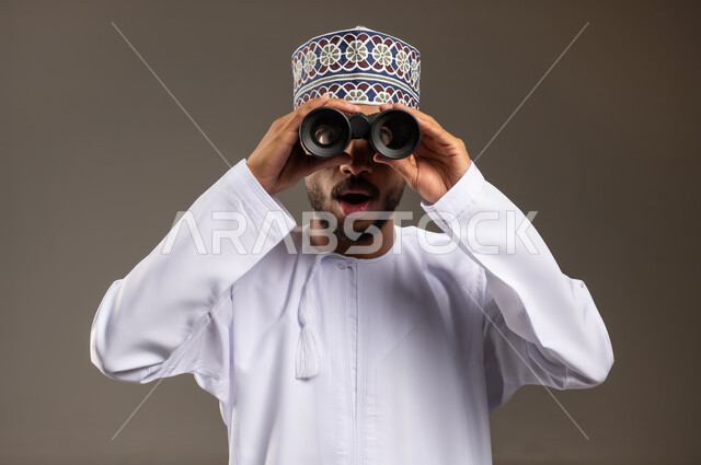 إيماءات الذهول والتفاجؤ, حب الاستكشاف و المغامرة, بورتريه مقرب لشاب عربي خليجي عماني مبتسم يرتدي الدشداشة و الكمة يحمل بيده منظار ثنائي العينين ينظر إلى شيئ ما, مفهوم النظرة المستقبلية، رؤية الاشياء البعيدة عبر التلسكوب, خلفية بيج