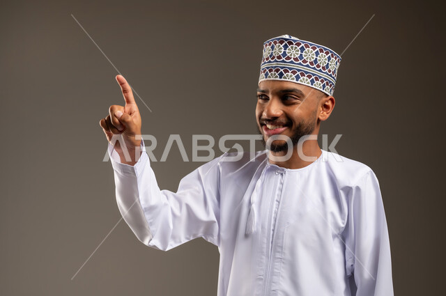 تحديد موقع معين بإيماءات السرور والبشاشة، بورتريه مقرب لرجل عربي خليجي عماني مبتسم يرتدي الدشداشة والكمة يشير بإصبع السبابة نحو الأعلى ينظر إلى شيء ما، إشارات وحركات يدين تدل على هذا المكان (هنا)، خلفية بيج