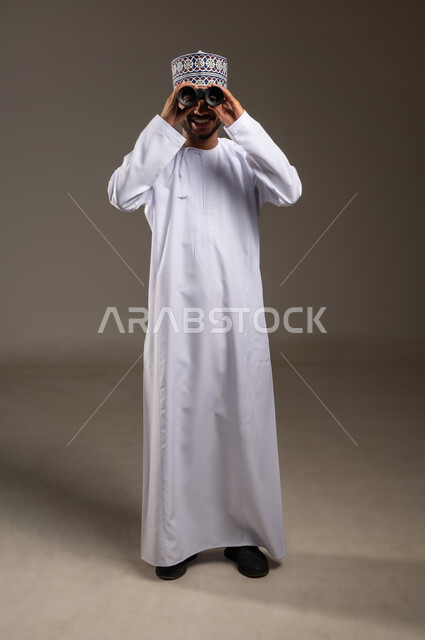 مفهوم النظرة المستقبلية، إيماءات الاستمتاع و السرور, حب الاستكشاف و المغامرة, بورتريه لشاب عربي خليجي عماني مبتسم يرتدي الدشداشة و الكمة يحمل بيده منظار ثنائي العينين ينظر إلى شيئ ما, رؤية الاشياء البعيدة عبر التلسكوب, صورة الجسم بالطول الكامل, خلفية بيج