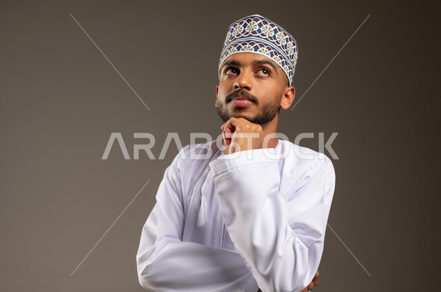 تعابير شرود الذهن والتمعن، إيجاد حل لمشكلة ما، بورتريه مقرب لرجل عربي خليجي عماني يرتدي الدشداشة والكمة يضع يده أسفل ذقنه ينظر إلى الأعلى بايماءات التفكير والتركيز، التأمل والتخطيط للمستقبل، خلفية بيج