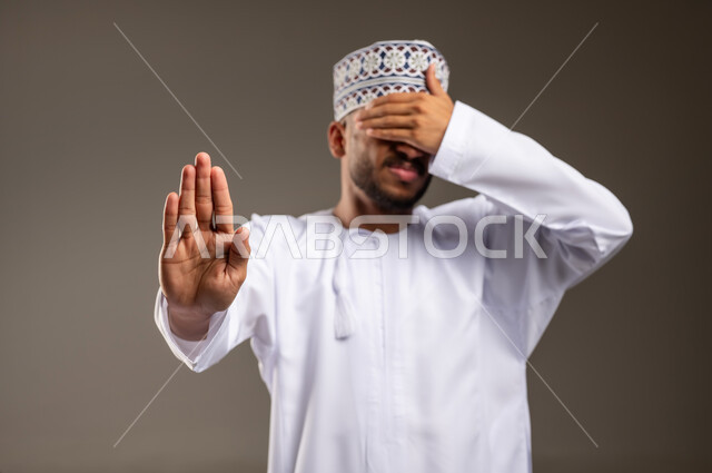 مفهوم الرفض والامتناع والانزعاج, بورتريه مقرب لرجل عربي خليجي عماني يرتدي الدشداشة والكمة يضع يده على عيونه يمد اليد الأخرى نحو الأمام مع رفع الكف بإيماءات النهي عن فعل امر ما, طلب التوقف وعدم الاقتراب, تعابير الحزم والجدية,  خلفية بيج