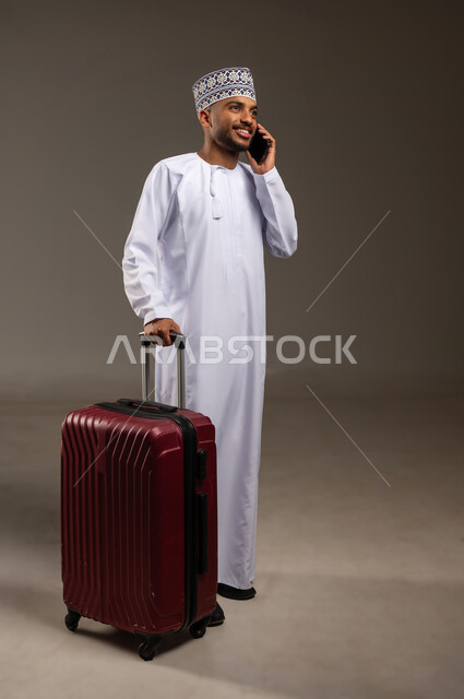 استخدام التقنيات الحديثة في التأكد من موعد الحجز, القيام بجولة ترفيهية، بورتريه لرجل عربي خليجي عماني يرتدي الدشداشة والكمة يقوم بجر حقيبة الأمتعة يجري مكالمة هاتفية عبر الهاتف المحمول، الفرح والاستعداد لقضاء رحلة صيفية ممتعة، الجسم بالطول الكامل، خلفية بيج