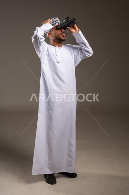 الذهول والتفاجؤ بمشاهدة أمر معين، رؤية الاشياء البعيدة عبر التلسكوب, حب الاستكشاف والمغامرة, بورتريه لشاب عربي خليجي عماني يرتدي الدشداشة والكمة يحمل بيده منظار ثنائي العينين يضع يده على رأسه ينظر إلى شيئ ما, صورة الجسم بالطول الكامل, خلفية بيج