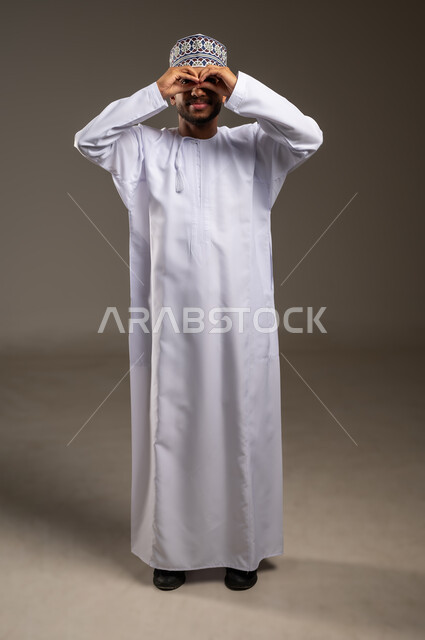 إيماءات التدقيق والتركيز، مشاهدة الاشياء الصغيرة بدقة ووضوح, حب الاستكشاف والمغامرة, بورتريه لشاب عربي خليجي عماني يرتدي الدشداشة والكمة يضع يديه على عينيه بشكل منظار، مفهوم توضيح الصورة، الجسم بالطول الكامل، خلفية بيج