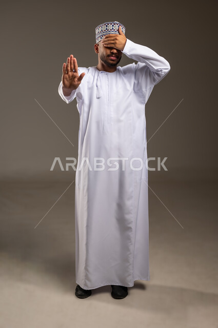 طلب التوقف وعدم الاقتراب, مفهوم الرفض والامتناع والانزعاج, بورتريه لرجل عربي خليجي عماني يرتدي الدشداشة والكمة يضع يده على عيونه يمد اليد الأخرى نحو الأمام مع رفع الكف بإيماءات النهي عن فعل امر ما, تعابير الحزم والجدية, صورة الجسم بالطول الكامل، خلفية بيج