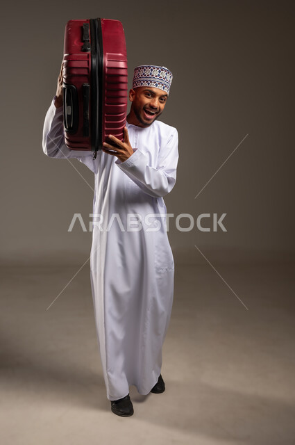 الفرح والاستعداد لقضاء رحلة صيفية ممتعة، مفهوم السياحة في إجازة الصيف، القيام بجولة ترفيهية، بورتريه لرجل عربي خليجي عماني يرتدي الدشداشة والكمة يحمل حقيبة الأمتعة على كتفه ينظر إلى الكاميرا بإيماءات السرور والحماس، الجسم بالطول الكامل، خلفية بيج
