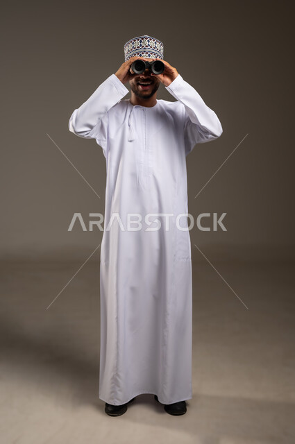 Seeing distant objects through a telescope, gestures of enjoyment and pleasure, love of exploration and adventure, portrait of a smiling young Omani Arab Gulf man wearing a dishdasha and a kuma holding binoculars in his hand looking at something, full-length body image, beige background