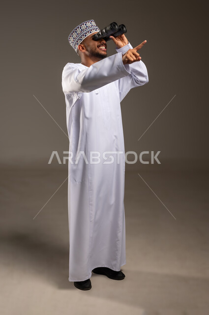 إيماءات الاستمتاع و السرور, حب الاستكشاف و المغامرة, بورتريه لشاب عربي خليجي عماني مبتسم يرتدي الدشداشة و الكمة يحمل بيده منظار ثنائي العينين يشير بإصبع السبابة للأعلى ينظر إلى شيئ ما, رؤية الاشياء البعيدة عبر التلسكوب, صورة الجسم بالطول الكامل, خلفية بيج