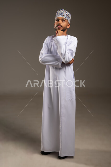 التأمل والتخطيط للمستقبل، تعابير شرود الذهن والتمعن، إيجاد حل لمشكلة ما، بورتريه لرجل عربي خليجي عماني يرتدي الدشداشة والكمة يضع يده أسفل ذقنه ينظر إلى الأعلى بايماءات التفكير والتركيز، الجسم بالطول الكامل، خلفية بيج