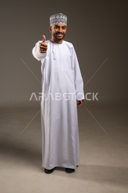 علامة الجودة والامتياز، حركات الإعجاب والقبول, الموافقة على أمر معين، بورتريه لرجل عربي خليجي عماني مبتسم يرتدي الدشداشة والكمة ينظر إلى الكاميرا يرفع إصبع الإبهام للأعلى بإيماءات الرضا والسرور والسعادة، صورة الجسم بالطول الكامل، خلفية بيج
