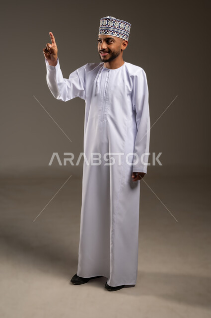 إشارات وحركات يدين تدل على هذا المكان (هنا)، تحديد موقع معين بإيماءات السرور والبشاشة، بورتريه لرجل عربي خليجي عماني مبتسم يرتدي الدشداشة والكمة يشير بإصبع السبابة نحو الأعلى ينظر إلى شيء ما، صورة الجسم بالطول الكامل، خلفية بيج