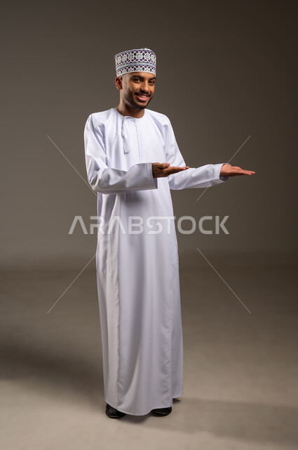 Determining a specific location or target, indicating the location of an item, portrait of a smiling Omani Gulf Arab man wearing a dishdasha and a kummah pointing to the palm of the other hand looking at the camera with gestures of pleasure, carrying and lifting something, concept of pointing and directing, full length body, beige background
