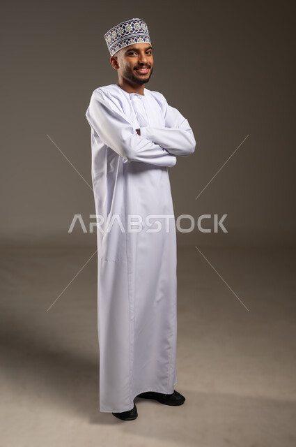 Concern for elegance and appearance, expressions of confidence and self-esteem, portrait of a smiling Arab Gulf Omani man wearing a dishdasha and a kummah standing with crossed hands looking at the camera with gestures of pleasure, concept of handsomeness and masculinity, full-length body, beige background
