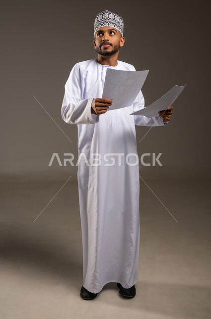 تدوين الملاحظات وإعداد التقارير اليومية، مراجعة نتائج الاجتماع، بورتريه لرجل عربي خليجي عماني يرتدي الدشداشة والكمة يحمل بيده مجموعة أوراق بيضاء ينظر إلى الأعلى بايماءات التفكير والتركيز، دراسة جدوى العمل، الجسم بالطول الكامل، خلفيه بيج