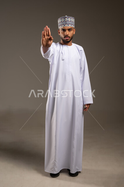 تعابير الحزم والجدية, طلب التوقف وعدم الاقتراب, مفهوم الرفض والامتناع والانزعاج, بورتريه لرجل عربي خليجي عماني يرتدي الدشداشة والكمه يمد يده نحو الأمام مع رفع الكف بإيماءات النهي عن فعل امر ما, صورة الجسم بالطول الكامل، خلفية بيضاء