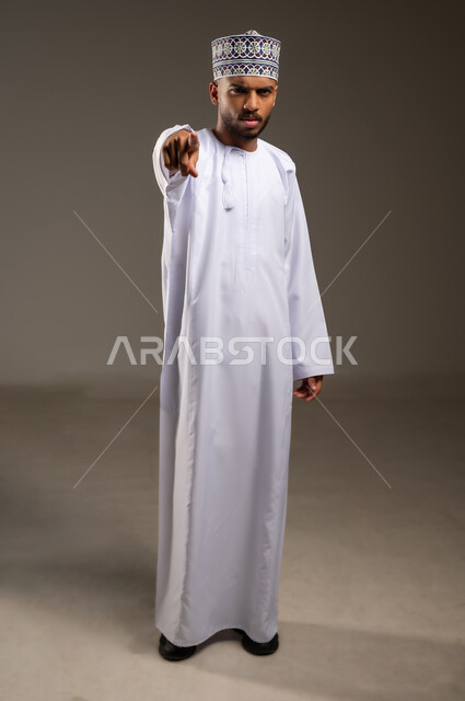 التوبيخ والاتهام وإلقاء اللوم، طلب التوقف عن فعل الأخطاء، الشعور بالانزعاج والرغبة بالانتقام، بورتريه لرجل عربي خليجي عماني يرتدي الدشداشة والكمة يرفع إصبع السبابة إلى الأمام لتحديد شخص ما ( أنت )، صورة الجسم بالطول الكامل، خلفية بيج
