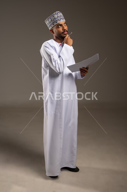 دراسة جدوى العمل، تدوين الملاحظات وإعداد التقارير اليومية، مراجعة نتائج الاجتماع، بورتريه لرجل عربي خليجي عماني يرتدي الدشداشة والكمة يحمل بيده مجموعة أوراق بيضاء وقلم يضع يده أسفل ذقنه بايماءات التفكير والتركيز، الجسم بالطول الكامل، خلفيه بيج