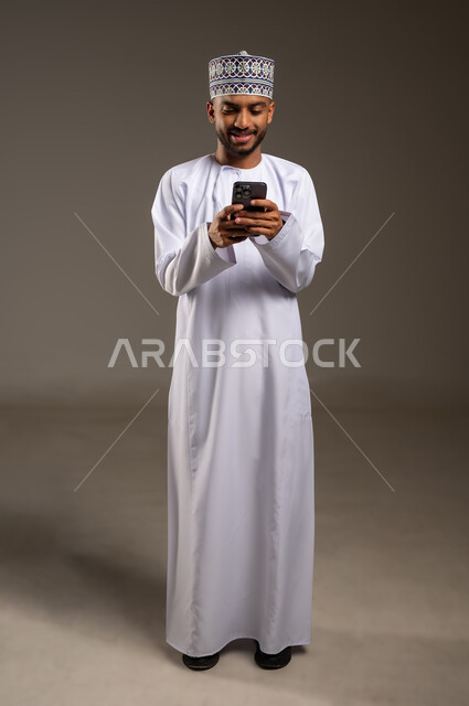 Browsing social media, chatting with family and friends, using a modern technical device, portrait of a young Arab Gulf Omani man wearing a dishdasha and a kummah holding a mobile phone, hearing good news from relatives, full body, beige background
