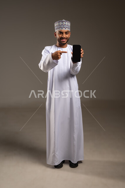 عرض شاشة سوداء فارغة، تصفح مواقع التواصل الاجتماعي, بورتريه لرجل عربي خليجي عماني يرتدي الدشداشة والكمة يحمل بيده الهاتف المحمول يشير إليه ينظر إلى الكاميرا بإيماءات السعادة والسرور, استخدام جهاز تقني حديث, الجسم بالطول الكامل، خلفية بيج