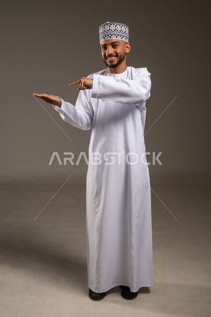 الدلالة على مكان عنصر، بورتريه لرجل عربي خليجي عماني مبتسم يرتدي الدشداشة والكمة يشير بإصبع السبابة إلى كف يده الأخرى ينظر إلى الكاميرا بإيماءات السرور، تحديد موقع أو هدف معين،  حمل ورفع شيء ما، مفهوم الإشارة والتوجيه، الجسم بالطول الكامل، خلفية بيج