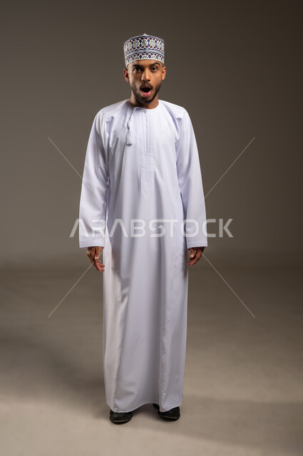 فتح الفم بإيماءات التعجب والتفاجؤ من شيء ما، تعابير الدهشة بسماع خبر مفاجئ، بورتريه لشاب عربي خليجي عماني يرتدي الدشداشة والكمة يقف باستقامة ينظر إلى الكاميرا، الاستغراب من أمر معين، صورة الجسم بالطول الكامل, خلفية بيج
