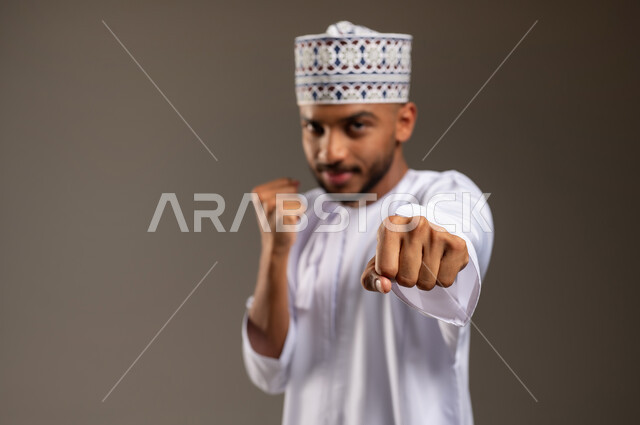 تعزيز صحة العظام والمفاصل، إتقان فن الملاكمة، بورتريه مقرب لرجل عربي خليجي عماني يرتدي الدشداشة التقليدية والكمة يرفع قبضات يديه ينظر إلى الكاميرا بإيماءات القوة، ممارسة رياضة الدفاع عن النفس، زيادة قدرة العضلات على التحمل، خلفية بيج
