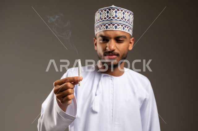 الأثر السلبي للإدمان على التبغ، المعاناة من الاضطرابات النفسية والمشاكل الصحية، بورتريه مقرب لرجل عربي خليجي عماني يرتدي الدشداشة والكمة يحمل بيده السيجارة ينظر إليها بإيماءات الكره والاشمئزاز، القيام بعادة سلوكية ضارة وتعاطي مادة سامة، خلفية بيج