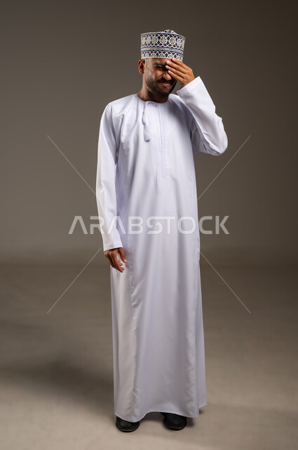 الاحساس بالشقيقية والصداع النصفي, الشعور بالإرهاق وفقدان التركيز, النسيان ومحاولة تذكر شيء ما, بورتريه لرجل عربي خليجي عماني يرتدي الدشداشة والكمة يضع يديه على رأسه ويغمض عينيه بإيماءات وجع في الدماغ والرأس الشديد, الجسم بالطول الكامل, خلفية بيج