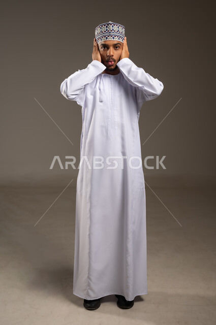 طلب السكوت والصمت، تعابير الدهشة بسماع خبر مفاجئ، بورتريه لشاب عربي خليجي عماني يرتدي الدشداشة والكمة يضع يديه على أذنيه ينظر إلى الكاميرا بإيماءات التعجب والتفاجؤ من شيء ما، صورة الجسم بالطول الكامل, خلفية بيج