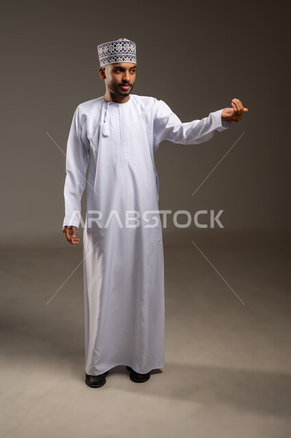 حمل ورفع شيء ما، تحديد موقع أو هدف معين، الدلالة على مكان عنصر، بورتريه لرجل عربي خليجي عماني يرتدي الدشداشة والكمة يرفع يده للأعلى، مفهوم الإشارة والتوجيه، الجسم بالطول الكامل، خلفية بيج