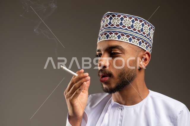 المعاناة من الاضطرابات النفسية والمشاكل الصحية، بورتريه مقرب لرجل عربي خليجي عماني يرتدي الدشداشة والكمة يقوم بتدخين السيجارة، القيام بعادة سلوكية ضارة وتعاطي مادة سامة، الأثر السلبي للإدمان على التبغ والمخدرات، خلفية بيج