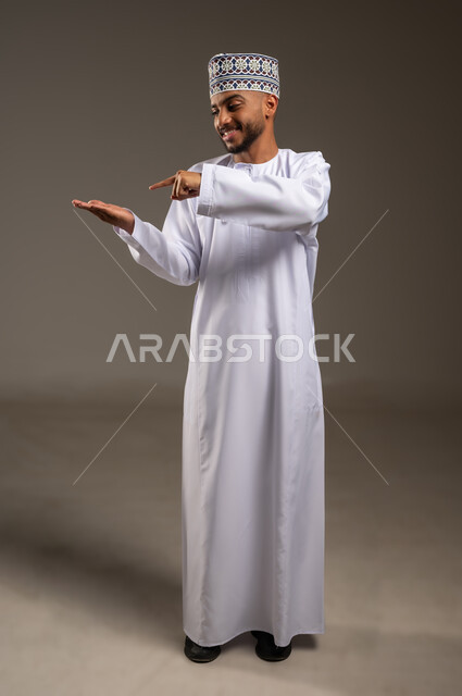 حمل ورفع شيء ما، الدلالة على مكان عنصر، بورتريه لرجل عربي خليجي عماني مبتسم يرتدي الدشداشة والكمة يشير بإصبع السبابة إلى كف يده الأخرى، تحديد موقع أو هدف معين، مفهوم الإشارة والتوجيه، الجسم بالطول الكامل، خلفية بيج