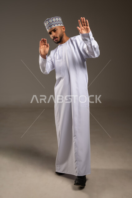 الذعر والخوف والصدمة، طلب التوقف والامتناع عن فعل شيء ما، بورتريه لرجل عربي خليجي عماني يرتدي الدشداشة التقليدية والكمة يرفع يديه للأعلى ينظر إلى الكاميرا بإيماءات الرعب وطلب النجدة، حماية النفس من ضرر خارجي, الجسم بالطول الكامل، خلفية بيج