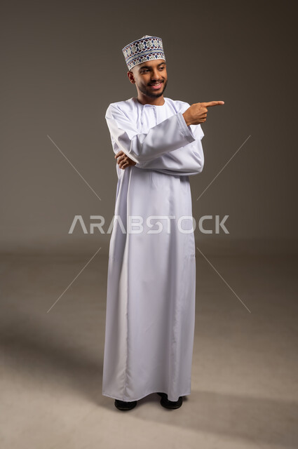 شد الإنتباه ولفت النظر، بورتريه لرجل عربي خليجي عماني مبتسم يرتدي الدشداشة والكمة يشير بإصبع السبابة ينظر إلى شيء ما، تحديد موقع أو هدف معين، الإشارة والتوجيه إلى مكان ما، الجسم بالطول الكامل، خلفية بيج