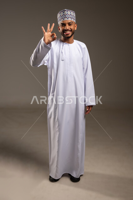 حركات الإعجاب والقبول, الموافقة على أمر معين، بورتريه لرجل عربي خليجي عماني مبتسم يرتدي الدشداشة والكمة ينظر إلى الكاميرا يرفع يده للأعلى بإيماءات علامة الجودة, تعابير الرضا والسرور والسعادة، صورة الجسم بالطول الكامل، خلفية بيج