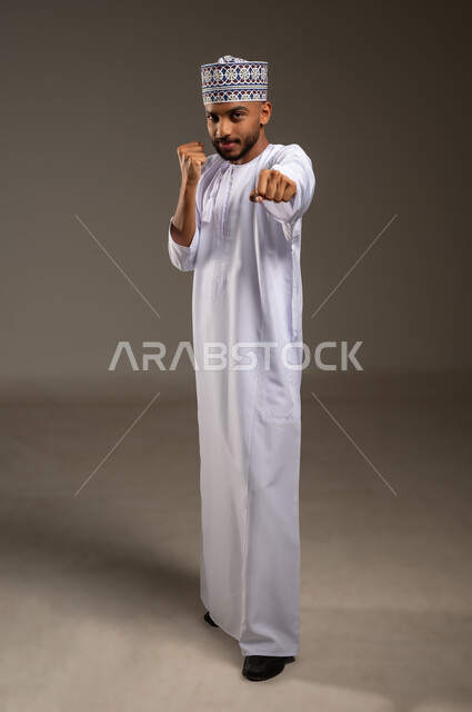 زيادة قدرة العضلات على التحمل، إتقان فن الملاكمة، بورتريه لرجل عربي خليجي عماني يرتدي الدشداشة التقليدية والكمة يرفع قبضات يديه ينظر إلى الكاميرا بإيماءات القوة، ممارسة رياضة الدفاع عن النفس، تعزيز صحة العظام والمفاصل، الجسم بالطول الكامل، خلفية بيج
