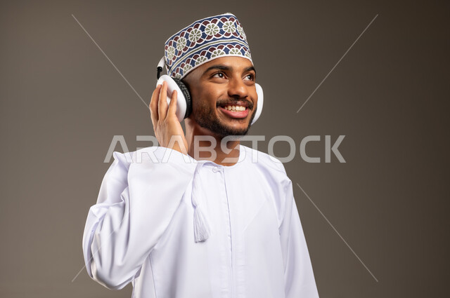 موهبة غنائية فنية، التفاعل مع الأنغام الموسيقية باستخدام الهيدفون، إيماءات الاندماج مع الأغنية، بورتريه مقرب لرجل عربي خليجي عماني يرتدي الدشداشة والكمة يستمع إلى الموسيقى من خلال سماعة الرأس بتعابير الانصات والاستمتاع، خلفية بيج