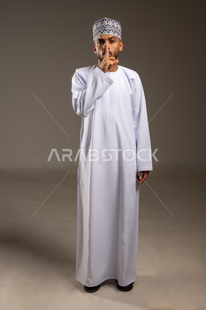 التوقف عن الكلام و الأمر بالصمت والسكوت، بورتريه لرجل عربي خليجي عماني يرتدي الدشداشة والكمة يضع إصبع السبابة على فمه ينظر إلى الكاميرا بإيماءات طلب الهدوء وعدم الازعاج, مفهوم الحاجة إلى التوقف والإنصات، الجسم بالطول الكامل، خلفية بيج