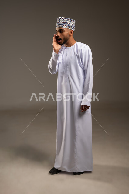 محادثات وتواصل اجتماعي، النداء بصوت مرتفع, وضع اليد بجانب الفم لزيادة حدة الأصوات، بورتريه من الجانب لشاب عربي خليجي عماني يرتدي الدشداشة والكمة ينظر إلى الكاميرا بإيماءات المناداة لشخص ما، الصراخ بنبرة عالية للسماع بشكل جيد، الجسم بالطول الكامل، خلفية بيج