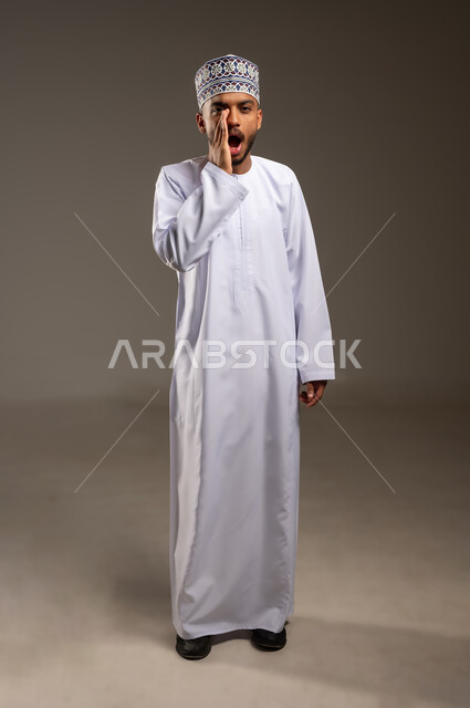 النداء بصوت مرتفع, وضع اليد بجانب الفم لزيادة حدة الأصوات، بورتريه لشاب عربي خليجي عماني يرتدي الدشداشة والكمة ينظر إلى الكاميرا بإيماءات المناداة لشخص ما، الصراخ والتكلم بنبرة عالية للسماع بشكل جيد، محادثات وتواصل اجتماعي، الجسم بالطول الكامل، خلفية بيج