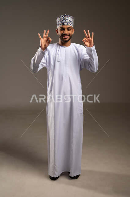 تعابير الرضا والسرور والسعادة، حركات الإعجاب والقبول, الموافقة على أمر معين، بورتريه لرجل عربي خليجي عماني مبتسم يرتدي الدشداشة والكمة ينظر إلى الكاميرا يرفع يديه للأعلى بإيماءات علامة الجودة, صورة الجسم بالطول الكامل، خلفية بيج