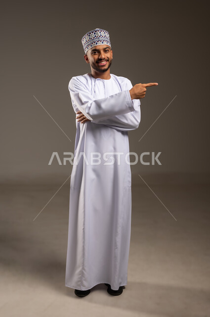 الإشارة والتوجيه إلى مكان ما، شد الإنتباه ولفت النظر، بورتريه لرجل عربي خليجي عماني مبتسم يرتدي الدشداشة والكمة يشير بإصبع السبابة إلى شيء ما ينظر إلى الكاميرا بإيماءات الثقة بالنفس، تحديد موقع أو هدف معين، الجسم بالطول الكامل، خلفية بيج