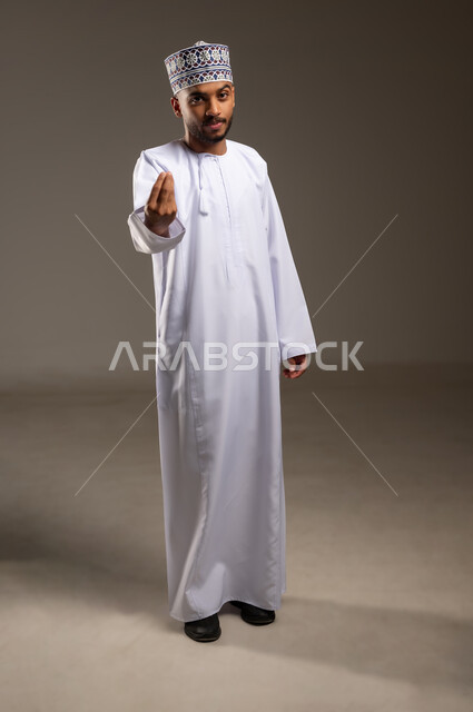 إمهال بعض الوقت للقيام بشئ ما، مفهوم الصبر والتريث، محاولة تهدئة نقاش حاد، بورتريه لشاب عربي خليجي عماني يرتدي الدشداشة والكمة يرفع يده ينظر إلى الكاميرا بإيماءات الانتظار، طلب خفض الصوت والهدوء بأدب واحترام، الجسم بالطول الكامل، خلفية بيج