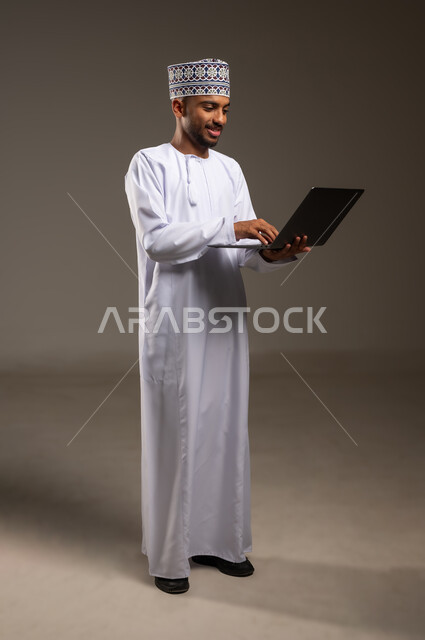 Using advanced modern technologies, managing and organizing work affairs, browsing social networking sites, portrait of an Arab Gulf Omani man wearing a dishdasha and a kimono working on a laptop with focus gestures, full body, beige background