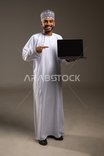 Using modern advanced devices and technologies, displaying a blank black screen, browsing social media sites, portrait of an Arab Gulf Omani man wearing a dishdasha and a kummah holding a laptop in his hand, pointing at it and looking at the camera with gestures of happiness and pleasure, full-length body, beige background