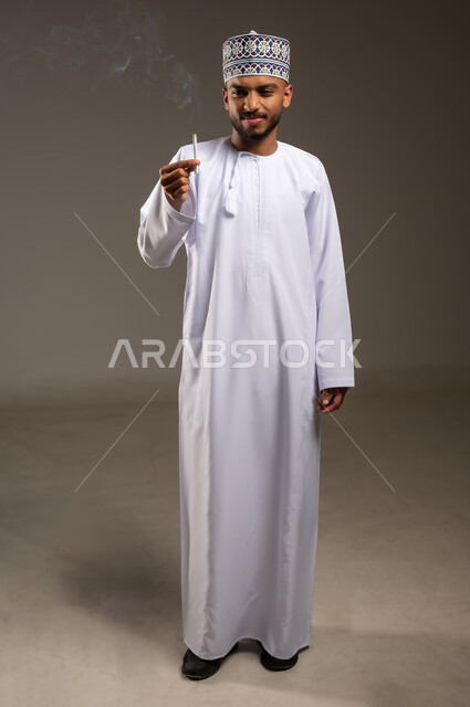 القيام بعادة سلوكية ضارة وتعاطي مادة سامة، الأثر السلبي للإدمان على التبغ، المعاناة من الاضطرابات النفسية والمشاكل الصحية، بورتريه لرجل عربي خليجي عماني يرتدي الدشداشة والكمة يحمل بيده السيجارة ينظر إليها بإيماءات الكره والاشمئزاز، الجسم بالطول الكامل، خلفية بيج