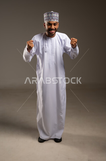 The concept of achievement and goal attainment, expressions of joy at winning and triumph, elegance, handsomeness and masculinity, portrait of a young Gulf Arab Omani man wearing a dishdasha and a kummah, raising his fists up, looking at the camera with gestures of pleasure and enthusiasm, full-length body, beige background
