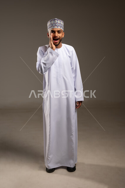 الصراخ والتكلم بنبرة عالية للسماع بشكل جيد، النداء بصوت مرتفع, وضع اليد بجانب الفم لزيادة حدة الأصوات، بورتريه لشاب عربي خليجي عماني يرتدي الدشداشة والكمة ينظر إلى الكاميرا بإيماءات المناداة لشخص ما، محادثات وتواصل اجتماعي، الجسم بالطول الكامل، خلفية بيج