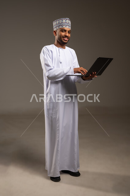 إدارة شؤون العمل وتنظيمه، تصفح مواقع التواصل الاجتماعي، بورتريه لرجل عربي خليجي عماني يرتدي الدشداشة والكمة يقوم بالعمل على الحاسوب المحمول بإيماءات التركيز ينظر إلى الكاميرا، استخدام التقنيات الحديثة المتطورة، الجسم بالطول الكامل، خلفية بيج